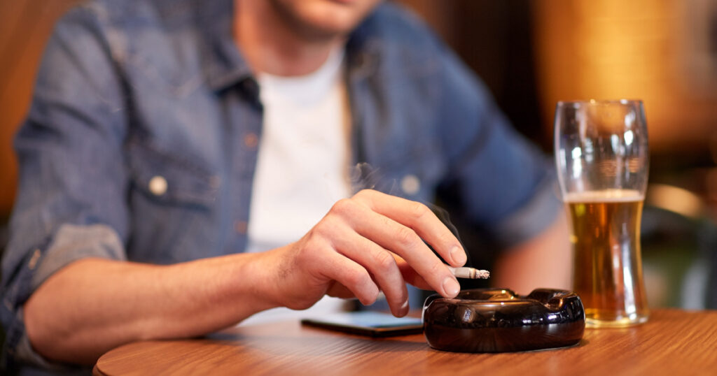 A person sitting at a table, holding a lit cigarette in one hand, with an ashtray nearby. A partially filled glass of beer is on the table. The person is wearing a denim jacket over a white shirt, and the background is blurred, focusing on the hand with the cigarette, the ashtray, and the glass of beer.
