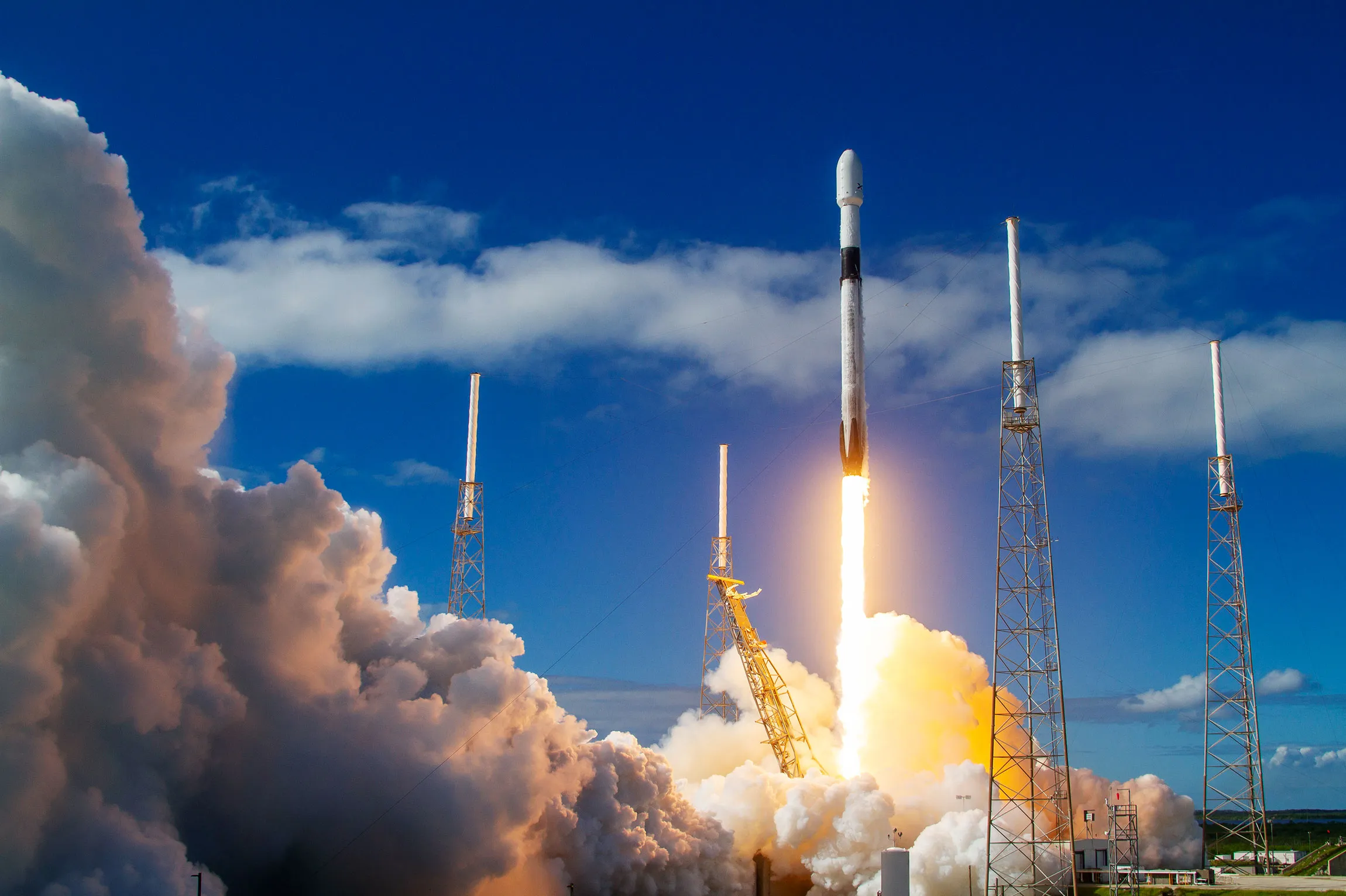 SpaceX Falcon 9 rocket launching into a clear blue sky with clouds of smoke billowing from the launch pad.