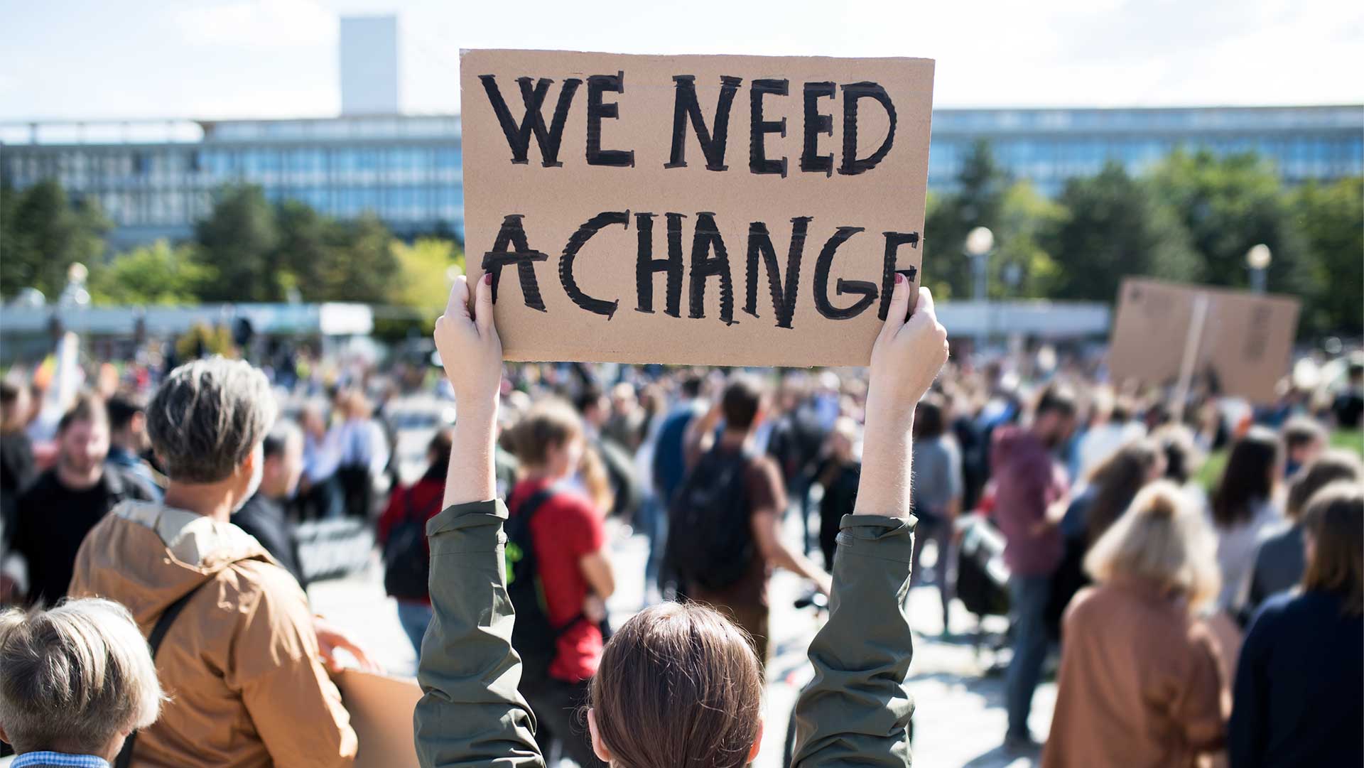 Person holding a sign that reads 'WE NEED A CHANGE' at a public protest with a large crowd in the background.