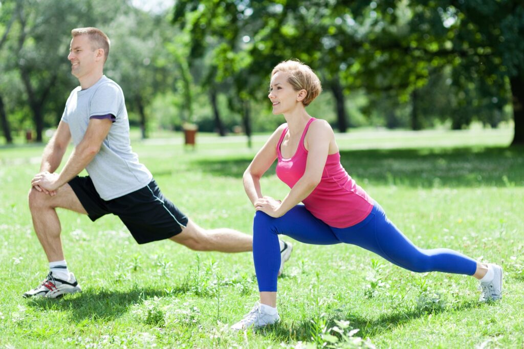 Two individuals performing a stretching exercise in an outdoor park. One person is wearing a gray t-shirt and black shorts, and the other is wearing a pink tank top and blue leggings. They are both in a lunge position with green grass and trees in the background.
